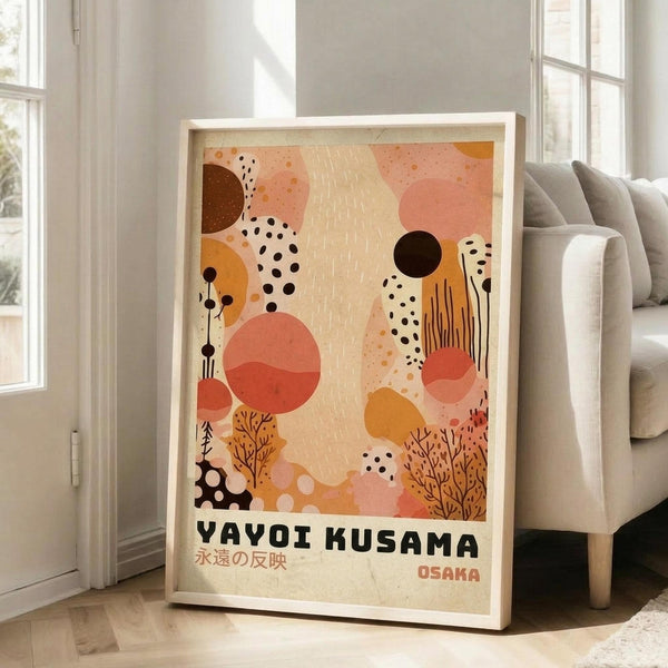 May include: Framed abstract art print leaning against white wall beside beige sofa in bright living room. Artwork features warm coral, orange, and brown organic shapes, dots, and botanical elements on cream background. Text reads "YAYOI KUSAMA" with Japanese characters and "OSAKA" below. Light wood frame, approximately 2-3 feet tall, positioned on light wooden floor near large windows.