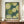 May include: Framed poster with abstract dot pattern artwork displayed on wooden credenza next to white ceramic vase and books. The print features overlapping circles in cream, olive green, and dark teal with polka dot patterns throughout. Text reads "YAYOI KUSAMA" with Japanese characters and "TOKYO" below. Warm lighting against cream colored paneled wall creates cozy interior setting.