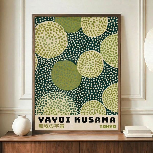 May include: Framed poster with abstract dot pattern artwork displayed on wooden credenza next to white ceramic vase and books. The print features overlapping circles in cream, olive green, and dark teal with polka dot patterns throughout. Text reads "YAYOI KUSAMA" with Japanese characters and "TOKYO" below. Warm lighting against cream colored paneled wall creates cozy interior setting.