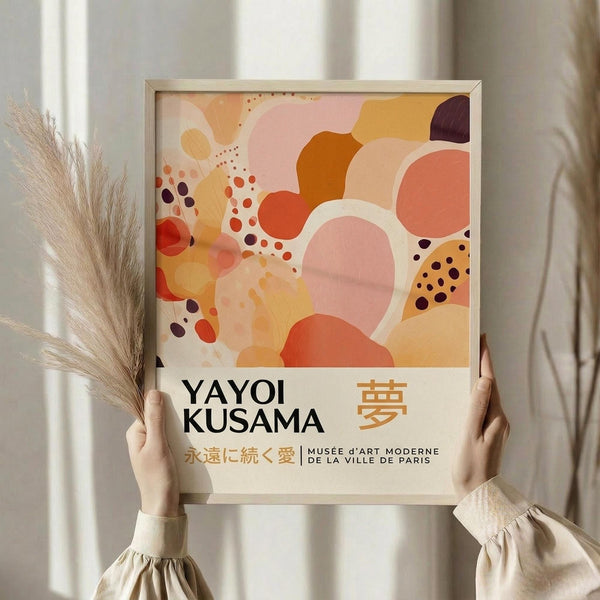 May include: Hands holding a framed art print featuring abstract organic shapes in warm pink, orange, coral and burgundy tones with scattered dots. The poster displays "YAYOI KUSAMA" in black text with Japanese characters and "MUSÉE d'ART MODERNE DE LA VILLE DE PARIS" below. Light wood frame against white curtained background with dried pampas grass visible to the left. Person wearing cream-colored ruffled sleeves.