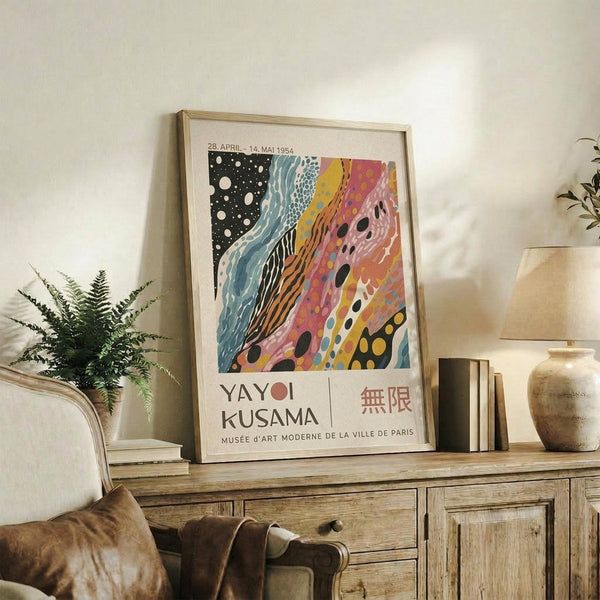 May include: Framed vintage-style exhibition poster with abstract colorful artwork featuring flowing patterns in black, blue, yellow, pink and orange, displayed on rustic wooden sideboard alongside fern plant, ceramic vases, and stacked books in neutral-toned living room setting