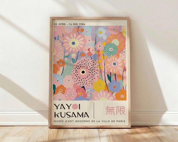 May include: Framed vintage exhibition poster featuring colorful abstract floral design with pastel pink, blue, orange and yellow blooms, polka dots and organic shapes. Text shows dates "28. APRIL - 14. MAI 1984" and "YAYOI KUSAMA" with "MUSÉE d'ART MODERNE DE LA VILLE DE PARIS" below. Light wood frame sits against white wall on blonde hardwood floor with natural window light creating soft shadows.