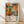 May include: Framed art print leaning against white wall on light wood floor. Colorful abstract botanical design with coral, orange, yellow and teal circular flowers, mushrooms and organic shapes in layered collage style. White mat border with "YAYOI KUSAMA" and "JAPAN 1987" text below artwork. Natural wood frame. Soft window light casting shadows across white textured wall.