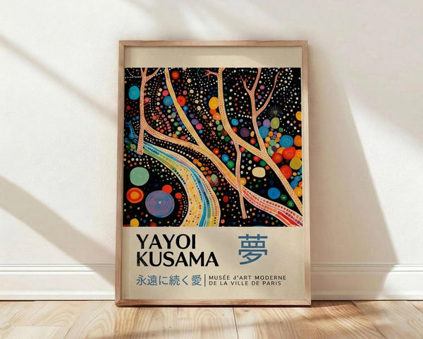 May include: Framed poster with abstract black background featuring colorful flowing organic shapes, dots, and circles in rainbow hues. Typography below artwork shows "YAYOI KUSAMA" and Japanese characters, plus "MUSÉE D'ART MODERNE DE LA VILLE DE PARIS". Natural wood frame sits against white wall on light wooden floor with soft shadows.