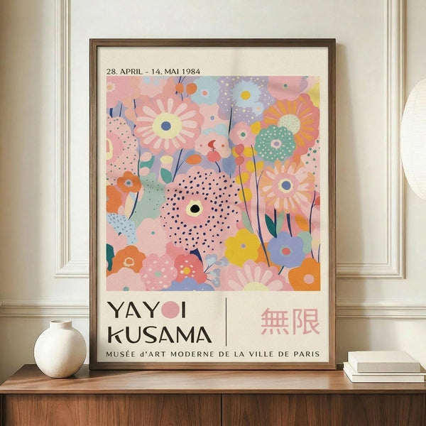 May include: Framed exhibition poster featuring colorful abstract floral design with pastel pink, blue, yellow and orange blooms, dots and patterns. Text shows dates "28. APRIL - 14. MAI 1984" and "YAYOI KUSAMA" with Japanese characters and "MUSÉE d'ART MODERNE DE LA VILLE DE PARIS". Brown wooden frame displayed on wooden sideboard next to white ceramic vase and stacked books against cream wall with white molding.
