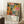 May include: Framed vintage-style art poster with colorful botanical illustration featuring abstract flowers, leaves, and coral forms in vibrant oranges, pinks, yellows and greens against dark background. Japanese text and "JAPAN 1987" visible at bottom. Poster leans against white wall on textured rug next to wooden side table with white coffee cup and saucer. Beige throw blanket partially visible on right side.
