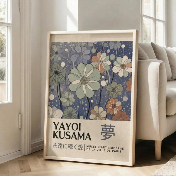 May include: Large framed art print leaning against white wall next to beige sofa in bright living room with windows. Print features stylized flowers in muted greens, peaches, and cream on deep blue dotted background, with Japanese text below including "YAYOI KUSAMA" and museum attribution. Light wooden frame, approximately 2-3 feet tall, positioned on light hardwood floor.