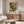 May include: Framed art print hanging above wooden credenza in modern living room setting. Print features colorful sunburst and dot pattern in green, orange, and cream tones with Japanese text. White sculptural chair and glass coffee table visible in foreground. Warm natural lighting illuminates the minimalist space with neutral walls.