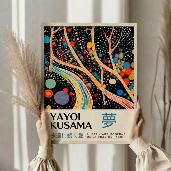 May include: Person holding framed poster with colorful abstract artwork featuring flowing ribbons and polka dots on black background. Poster displays "YAYOI KUSAMA" text and Japanese characters below vibrant dotted design with swirling lines in rainbow colors. Light wood frame against neutral wall with decorative dried pampas grass visible on left side.