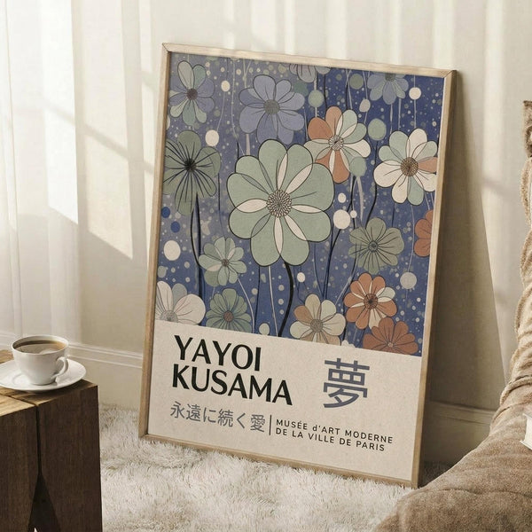May include: Framed art print leaning against white wall featuring stylized flowers in muted green, coral, and white tones against deep blue dotted background, with Japanese text below. Natural lighting creates soft shadows. White coffee cup on wooden surface and textured throw blanket visible nearby in cozy interior setting.