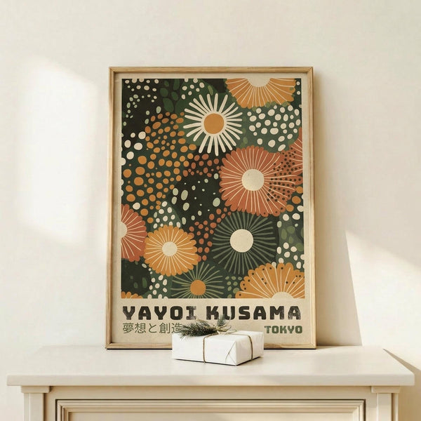 May include: Framed art print with colorful abstract floral design featuring radiating sunburst patterns in orange, green, and cream tones against dark background, displayed on light wood console table next to small wrapped gift with evergreen sprig, against white wall in minimalist interior setting.