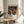 May include: Framed art print displayed on light wood round side table next to beige upholstered chair. Artwork features black background with vibrant multicolored dots and flowing tree-like branches in orange, blue, yellow, red, and green. White text below reads "YAYOI KUSAMA" with Japanese characters and museum attribution. Natural lighting creates soft shadows on white wall behind.