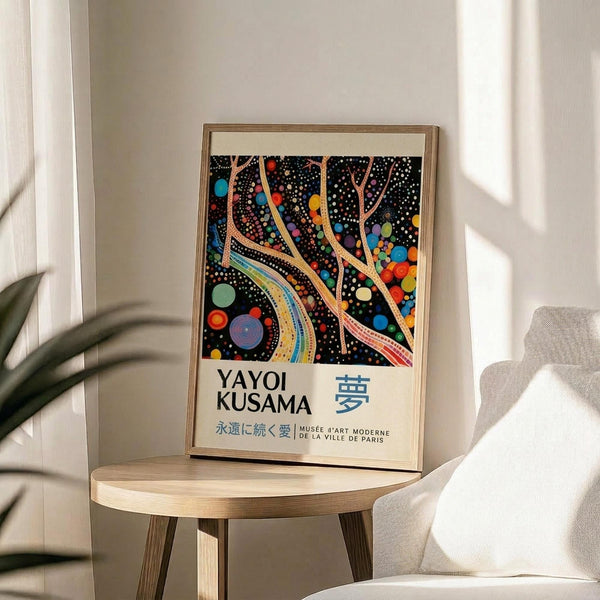 May include: Framed art print displayed on light wood round side table next to beige upholstered chair. Artwork features black background with vibrant multicolored dots and flowing tree-like branches in orange, blue, yellow, red, and green. White text below reads "YAYOI KUSAMA" with Japanese characters and museum attribution. Natural lighting creates soft shadows on white wall behind.