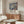 May include: Framed floral art print with blue background and colorful cosmos flowers displayed on wooden sideboard in modern living room. White curved chair and glass coffee table visible. Print features Japanese text and museum attribution. Warm natural lighting creates shadows on light colored walls.