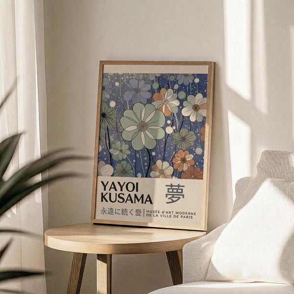May include: Framed poster with blue background featuring stylized flowers in green, orange, and cream tones with white dots, displayed on round wooden side table next to beige upholstered chair. Natural lighting creates soft shadows on neutral wall. Plant leaves visible in corner. Poster shows Japanese text and museum attribution at bottom.
