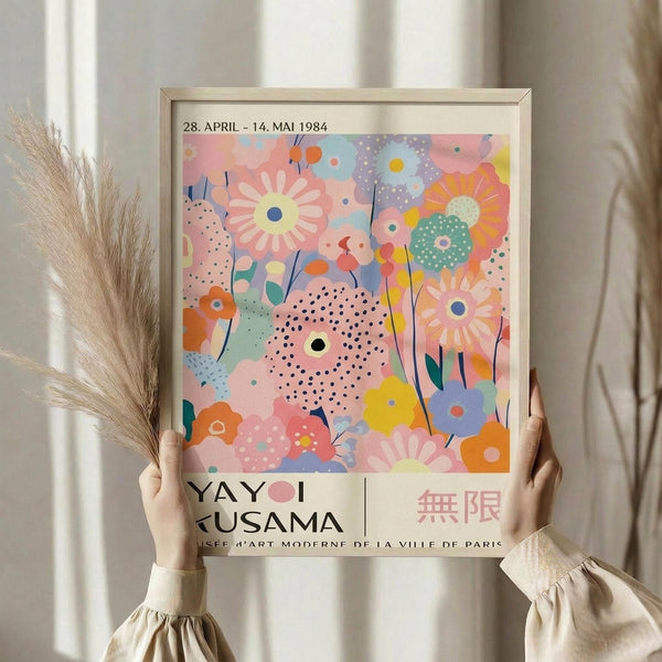 May include: Person holding framed art print featuring colorful abstract floral design with polka dots in pink, blue, yellow, and orange tones. Shows exhibition text "28. APRIL - 14. MAI 1984" and "YAYOI KUSAMA" at bottom. Light wood frame against neutral curtained background, with dried wheat stems visible on left side.