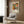 May include: Colorful Yayoi Kusama exhibition poster featuring vibrant coral, orange, and green organic forms mounted in light wood frame on textured brown wall. Modern beige armchair with geometric patterned pillow sits below artwork next to brass floor lamp and dark round side table with open book.