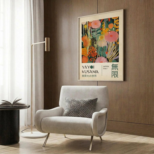 May include: Colorful Yayoi Kusama exhibition poster featuring vibrant coral, orange, and green organic forms mounted in light wood frame on textured brown wall. Modern beige armchair with geometric patterned pillow sits below artwork next to brass floor lamp and dark round side table with open book.