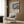 May include: Framed floral art print with blue background and colorful flowers in cream and earth tones, displayed on wood paneled wall above beige upholstered armchair with geometric throw pillow. Modern brass floor lamp and dark round side table with open book visible. Warm, contemporary living room setting with natural lighting from window.