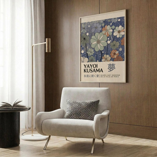 May include: Framed floral art print with blue background and colorful flowers in cream and earth tones, displayed on wood paneled wall above beige upholstered armchair with geometric throw pillow. Modern brass floor lamp and dark round side table with open book visible. Warm, contemporary living room setting with natural lighting from window.