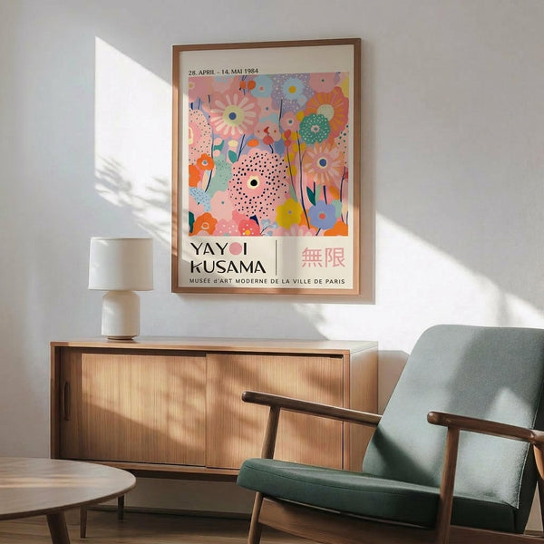 May include: Colorful Yayoi Kusama exhibition poster in wooden frame hanging above mid-century modern furniture. The artwork features vibrant floral patterns in pink, orange, green, and blue with polka dots. Room shows sage green upholstered chair with wooden arms, natural wood sideboard, white ceramic lamp, and warm sunlight casting shadows on white walls.