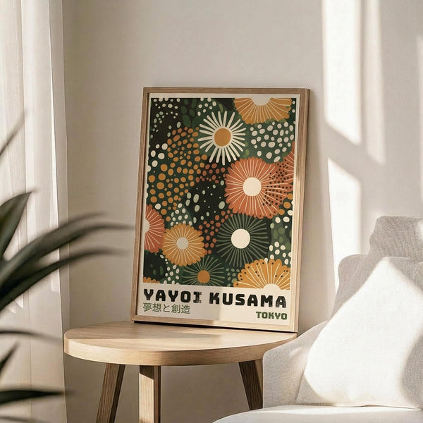 May include: Framed poster with colorful abstract floral pattern in green, orange, and cream tones featuring radiating sunburst shapes and polka dots, displayed on round wooden side table next to beige upholstered chair with plant leaves visible in bright, naturally lit room with white walls