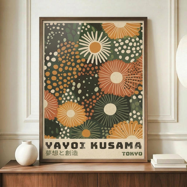 May include: Framed art print displayed on wooden sideboard featuring abstract floral design with radiating daisy-like flowers in orange, green, and cream colors against dark background with scattered dots. Japanese text below reads "Yayoi Kusama" and includes Tokyo reference. White ceramic vase and books sit nearby on warm wood surface against cream wall with white trim molding.