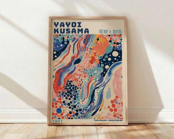 May include: Framed poster print leaning against white wall on light wooden floor. Light wooden frame contains colorful abstract artwork with flowing organic shapes, polka dots, and swirling patterns in navy blue, coral orange, teal, and cream. Text reads "YAYOI KUSAMA" with Japanese characters and "1991" at bottom. Natural sunlight creates soft shadows on white textured wall behind frame.