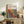 May include: Framed poster with Yayoi Kusama's name and Japanese text at top, featuring colorful abstract polka dot pattern in red, orange, yellow, and blue sections with black oval shapes throughout. Light wood frame displayed on rustic wooden sideboard alongside fern plant, books, ceramic vases, and cream upholstered chair in neutral-toned living room setting.