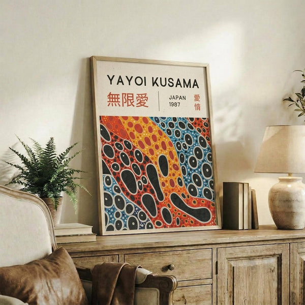 May include: Framed poster with Yayoi Kusama's name and Japanese text at top, featuring colorful abstract polka dot pattern in red, orange, yellow, and blue sections with black oval shapes throughout. Light wood frame displayed on rustic wooden sideboard alongside fern plant, books, ceramic vases, and cream upholstered chair in neutral-toned living room setting.