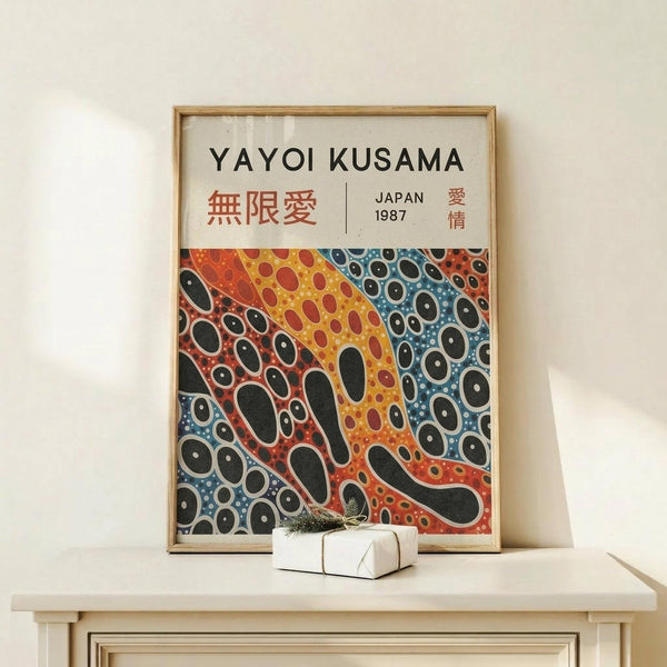 May include: Framed poster displaying colorful abstract artwork with organic flowing shapes and polka dot patterns in red, orange, yellow, and blue tones. Features text "YAYOI KUSAMA" and Japanese characters with "JAPAN 1987". Natural wood frame sits on light wooden surface next to small white wrapped gift with green sprig against cream wall.