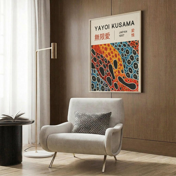 May include: Colorful Yayoi Kusama exhibition poster with polka dot pattern in orange, blue, and black tones, displayed in white frame on wood-paneled wall above beige upholstered armchair with geometric pillow, brass floor lamp and side table with open book visible in modern living room setting
