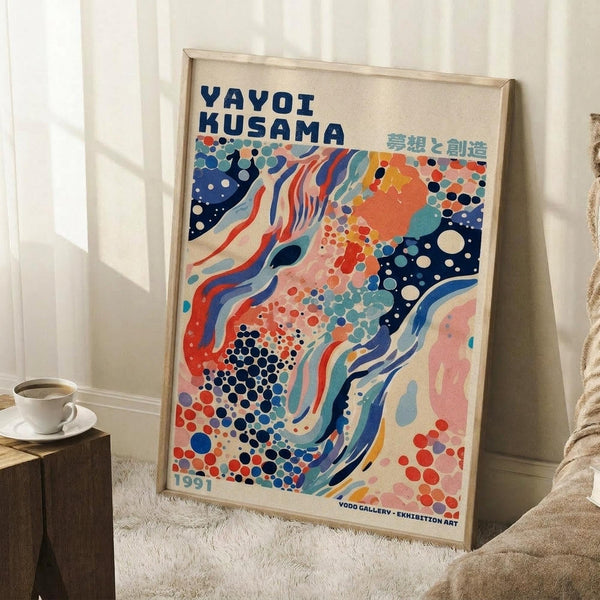 May include: Framed art poster with abstract colorful design featuring flowing organic shapes in navy blue, coral red, teal, and cream with polka dot patterns. Text reads "YAYOI KUSAMA" at top and "1991" at bottom. Poster leans against white wall next to wooden side table with white coffee cup, positioned on light textured rug near beige upholstered furniture in bright room with natural lighting.