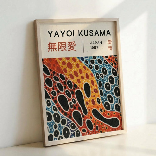 May include: Framed art print leaning against white wall featuring Yayoi Kusama's signature polka dot pattern in vibrant orange, yellow, blue and red organic shapes with black oval spots throughout. Text reads "YAYOI KUSAMA" at top with Japanese characters and "JAPAN 1987" below. Natural wood frame, white matting, positioned on light surface with soft shadows.