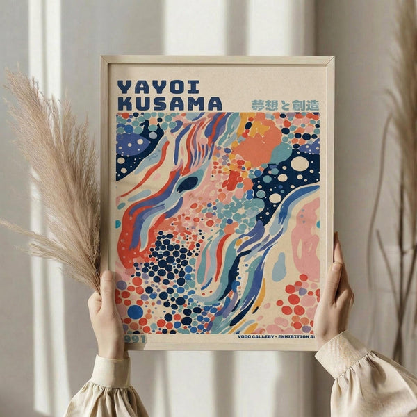 May include: Hands holding a framed colorful abstract art poster featuring flowing organic shapes in coral, navy, teal and cream with polka dot patterns. The poster displays "YAYOI KUSAMA" text at top with Japanese characters. Light wood frame against neutral curtained background with dried wheat stems visible on left side. Person wearing cream colored ruffled sleeve top.