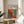 May include: Framed poster featuring abstract pattern of colorful dots and organic shapes in orange, blue, and black tones, displayed on light wood round side table next to beige upholstered chair. Natural lighting creates soft shadows on white wall background with green plant leaves visible in corner.
