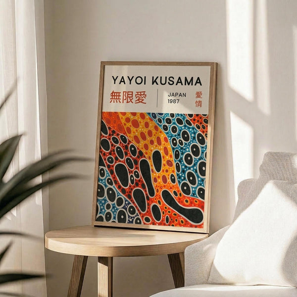 May include: Framed poster featuring abstract pattern of colorful dots and organic shapes in orange, blue, and black tones, displayed on light wood round side table next to beige upholstered chair. Natural lighting creates soft shadows on white wall background with green plant leaves visible in corner.