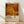 May include: Framed poster print leaning against white wall on light wood floor. Features vibrant orange, yellow, and brown polka dots in various sizes creating abstract pattern. Bottom shows cream-colored section with "YAYOI KUSAMA" text and Japanese characters, plus museum credit line. Natural wood frame with warm lighting casting soft shadows on textured white wall.