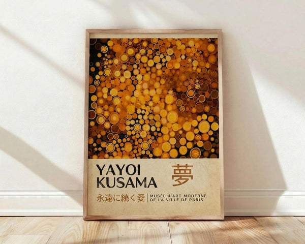 May include: Framed poster print leaning against white wall on light wood floor. Features vibrant orange, yellow, and brown polka dots in various sizes creating abstract pattern. Bottom shows cream-colored section with "YAYOI KUSAMA" text and Japanese characters, plus museum credit line. Natural wood frame with warm lighting casting soft shadows on textured white wall.