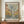 May include: Framed museum poster featuring abstract tree design with circular dots and organic shapes in muted green, yellow, blue and brown tones, displayed on wooden sideboard next to white ceramic vase and books in modern interior with white paneled walls
