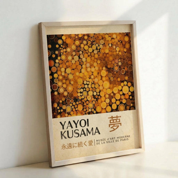 May include: Framed poster displayed against white wall featuring abstract pattern of golden, amber, and brown circular dots in various sizes creating polka dot design. Text shows "YAYOI KUSAMA" with Japanese characters and "MUSÉE D'ART MODERNE DE LA VILLE DE PARIS" on cream background. Light wooden frame with natural wood grain visible, casting subtle shadow on wall.