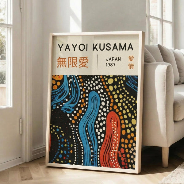 May include: Framed art print leaning against white wall next to beige sofa in bright living room. Poster features flowing blue and red organic shapes with white and multicolored polka dots on black background. Text reads "YAYOI KUSAMA" with Japanese characters and "JAPAN 1987". Light wood frame, approximately 2-3 feet tall, positioned on hardwood floor near large windows.