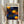 May include: Person in beige ruffled sleeves holding framed art print in light wooden frame. Artwork features vibrant blue background with orange and yellow polka dots in various sizes, three prominent orange spiral circles resembling suns or flowers. Japanese text visible at top. Dried pampas grass stems held in left hand against neutral curtained background.