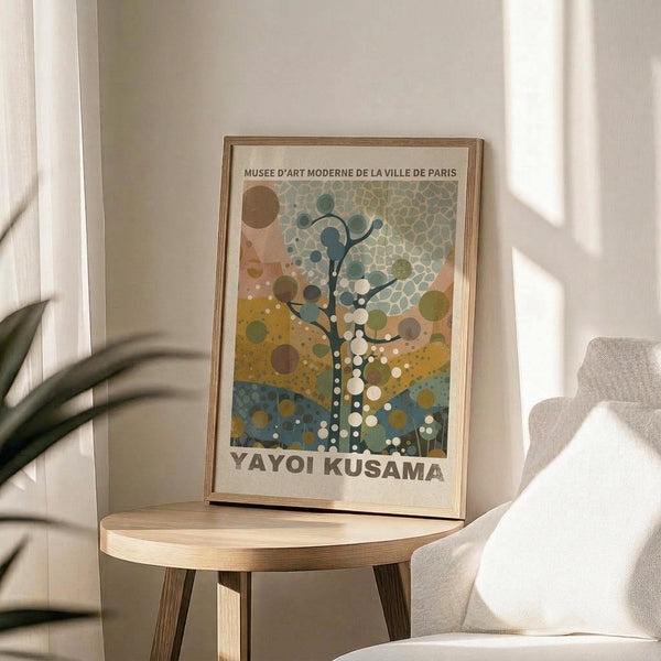 May include: A framed art print featuring a stylized tree with circular dots in earth tones of blue, green, yellow and brown, displayed on a wooden round side table next to a beige upholstered chair. Natural sunlight streams across the neutral-toned room setting with a plant visible in the corner.