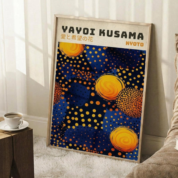 May include: Framed poster with blue background featuring yellow and orange polka dots in various sizes, three prominent yellow circular swirl motifs, and black text "YAYOI KUSAMA" with Japanese characters and "KYOTO" in orange. Light wood frame leaning against white wall corner next to white coffee cup on wooden block and cream textured blanket on white fluffy rug.