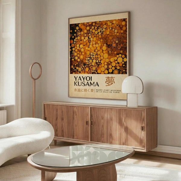 May include: Modern living room with wooden sideboard displaying framed Yayoi Kusama exhibition poster featuring golden and amber polka dot pattern against cream background. White sculptural chair, glass oval coffee table, and minimalist lamp complete the warm, contemporary interior scene.