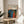 May include: Framed poster with abstract art in blue, red, orange and white dotted patterns reminiscent of flowing organic forms, displayed on round wooden side table next to beige upholstered chair. Natural lighting creates soft shadows on white wall background, with plant leaves visible in corner of minimalist modern interior setting.