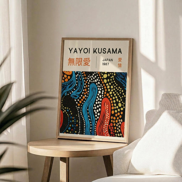 May include: Framed poster with abstract art in blue, red, orange and white dotted patterns reminiscent of flowing organic forms, displayed on round wooden side table next to beige upholstered chair. Natural lighting creates soft shadows on white wall background, with plant leaves visible in corner of minimalist modern interior setting.