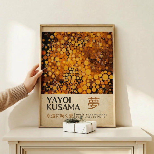 May include: Framed art print featuring orange and amber circular dots in Kusama's signature polka dot style against dark background, with artist name and Japanese characters below. Person in beige sweater holds wooden-framed poster on light wood surface next to small white wrapped gift with greenery. Cream colored wall background.
