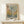 May include: Framed poster in natural wood frame leaning against white wall on light wooden surface. Features abstract tree design with circular dots in muted greens, blues, and earth tones. Text reads "Musee d'Art Moderne de la Ville de Paris" and "Yayoi Kusama" at bottom. Small white wrapped gift with green sprig sits beside frame. Minimalist room setting with neutral colors.